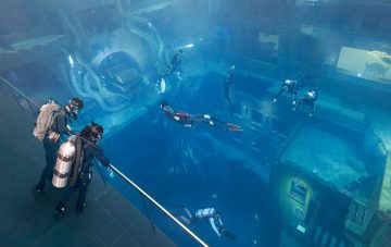 Wide overhead view of multiple scuba divers and freedivers exploring different levels of a submerged urban landscape.