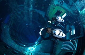 Two scuba divers hover above a deep, dark circular shaft surrounded by submerged buildings and a motorcycle.