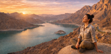 Woman sitting peacefully on a cliff overlooking Hatta Dam with mountains and lake at sunset