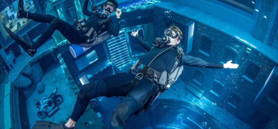 Two scuba divers using underwater lights to explore a room with exposed pipes, a workbench, and brick archways.