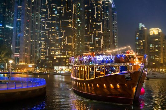 A traditional wooden dhow boat illuminated with gold lights cruising through a modern marina at night, surrounded by glowing skyscrapers.