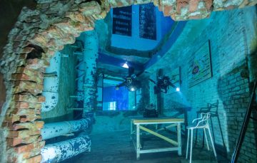 Two scuba divers swim through a room with exposed white pipes, a wooden table, and brick archways.