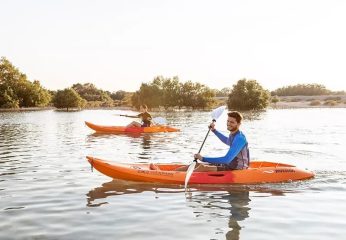 Two people in orange kayaks paddling across calm water during a bright, misty sunrise.