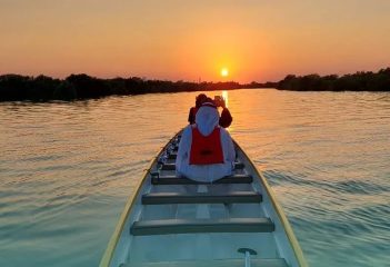 The interior view of a long white dragon boat heading toward a vibrant orange sunset over the water.