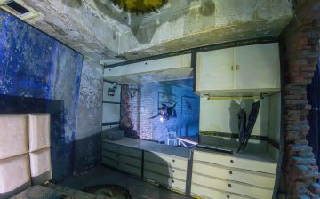 A scuba diver with a flashlight explores a fully furnished submerged bedroom with a bed, dresser, and wardrobe.