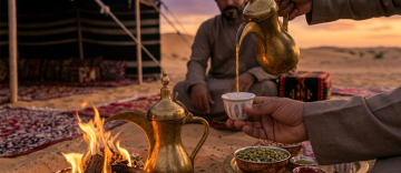 Bedouin host pouring Arabic coffee from a brass dallah into a finjan beside a desert campfire at sunset in Abu Dhabi