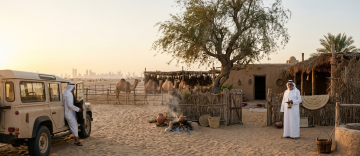 Traditional Ezba desert home with camels, campfire, and Emirati man holding dallah coffee pot