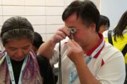 Person inspecting a gemstone with a loupe at a diamond exchange event, with a woman beside holding a mobile device.