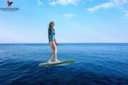 A woman gracefully balancing on an e-Foil board over the ocean under a bright blue sky.