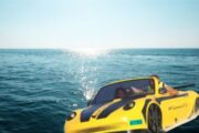 A man flexing confidently while standing on a yellow jet car parked on the water.