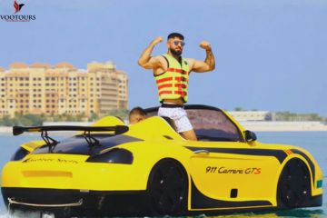 A man flexing confidently while standing on a yellow jet car parked on the water.