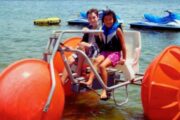 Two kids on an orange aqua cycle having fun near jet skis and water toys