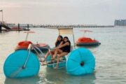 wo women on a blue aqua cycle in shallow waters, with orange pedal boats in the background