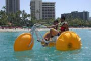 A couple enjoying a ride on a yellow water trike in a tropical setting with a hotel in the background