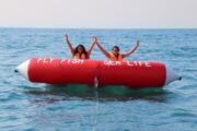 Two participants raising hands on a Fly Fish inflatable in the sea.