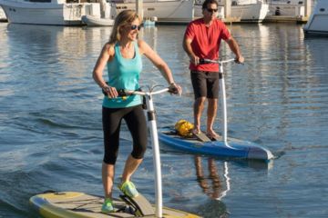 A man and woman riding Electric Stand-Up Paddle Boards near a marina.