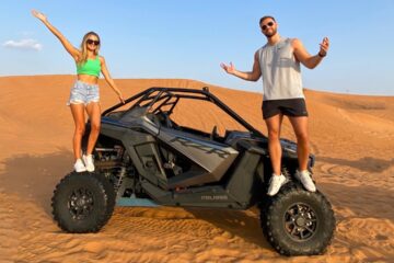 Couple standing on a dune buggy in the middle of the desert.