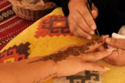 A woman receiving an intricate henna design on her hand at a desert camp.