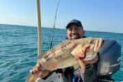 Angler holding a large grouper on a fishing boat with a backdrop of the open sea.