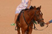 A young girl waving happily while riding a brown horse in the desert.