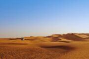 White 4×4 vehicle driving across golden desert dunes under a clear blue sky.