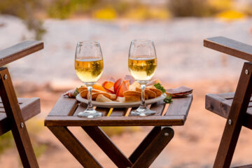 Two glasses of white wine and a plate of fruit on a small wooden table in a desert setting.