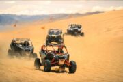 Multiple CAN-AM Maverick buggies racing across the sand dunes.