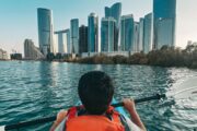 A child in an orange life vest kayaks toward Abu Dhabi’s skyline on calm waters.