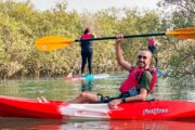 A smiling kayaker in a red kayak lifts his paddle overhead near green mangroves.