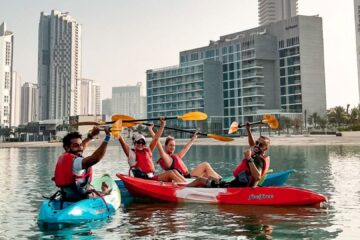 Four kayakers in brightly colored kayaks raise their paddles in celebration against a city skyline.