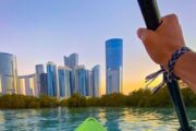 A kayaker’s hand grips a paddle, aiming toward Abu Dhabi’s skyscrapers under a colorful sky.