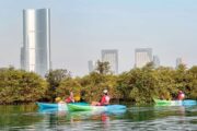 Three kayakers wearing life jackets paddle near lush mangroves with Abu Dhabi’s modern skyline in the background.