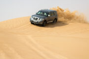 A 4x4 vehicle speeding over sand dunes, kicking up golden sand.