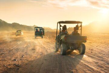 A convoy of off-road vehicles driving across dusty desert terrain with mountains in the background.