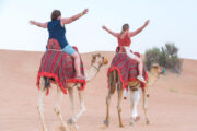 Tourists riding camels in the desert, led by a guide.