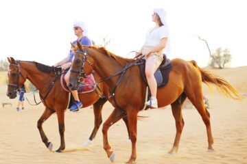 A young girl waving happily while riding a brown horse in the desert.