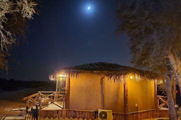 A circular, thatched-roof hut illuminated under a crescent moon in the desert.