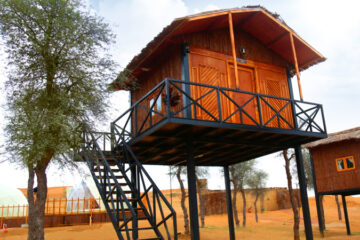 An elevated wooden cabin on stilts with a small balcony, set in a desert camp.