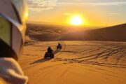 Two quad bikers riding across golden desert dunes at sunrise.