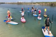 A cheerful group of stand-up paddleboarders posing on their boards in shallow, clear water with a cityscape in the background.