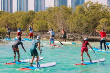 A group of stand-up paddleboarders navigating turquoise waters beside mangroves with a modern city skyline in the distance.