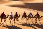 A caravan of camels with riders trekking across vast, golden desert dunes.