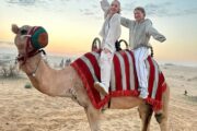 caravan of camels with riders trekking across vast, golden desert dunes.