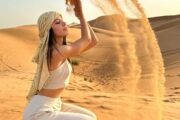 Woman in a white outfit and headscarf tossing sand into the air in a desert.