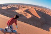 A woman standing on a sand board in a sunlit desert, poised for her next descent.