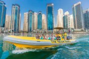 A guide speaking to passengers on a bright yellow boat with Dubai’s skyline in the background.