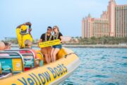 A smiling couple holding a “Wish You Were Here?” sign on a yellow boat, with Atlantis the Palm in the background.