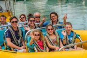 A smiling family group wearing life jackets, seated on a bright yellow boat, waving at the camera.