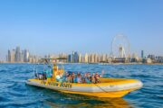 A bright yellow tour boat cruising off the Dubai coast with the city skyline and Ain Dubai in the background.