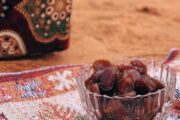 A small Arabic coffee cup and a bowl of dates arranged on a patterned mat in the desert.